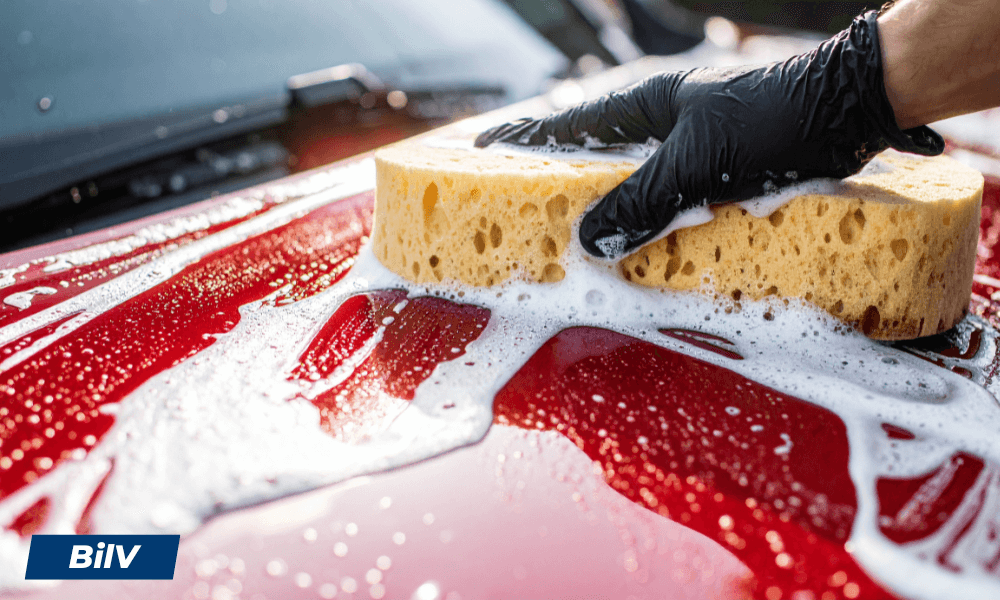 Hand med handske tvättar bilens motorhuv med gul bilsvamp och skum.